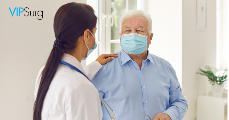 Female Doctor hand on patient and talking to an older male patient with a mask on in a blue button down shirt